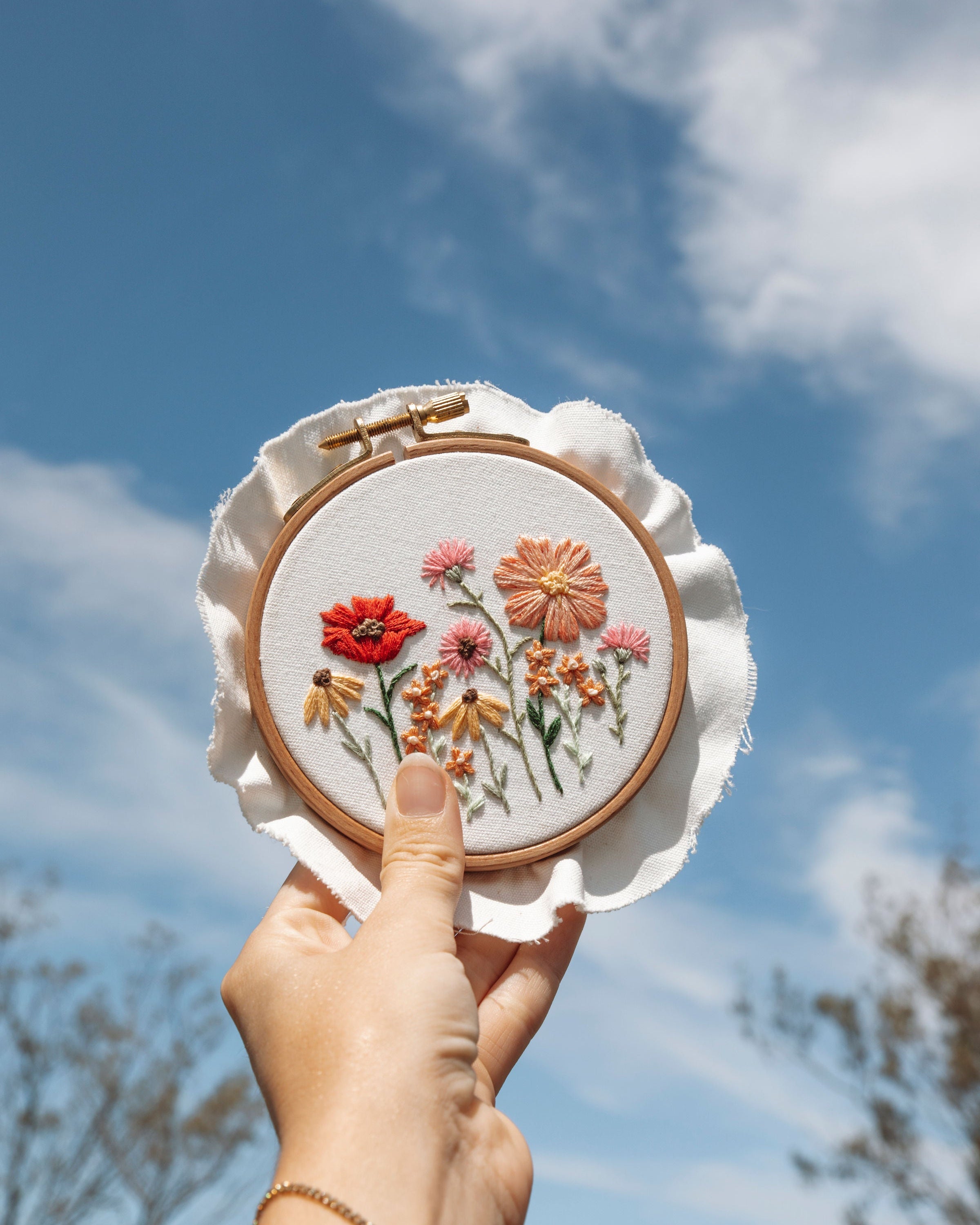 Hand holding embroidery hoop with colorful stitched wildflowers against a bright blue sky with soft clouds