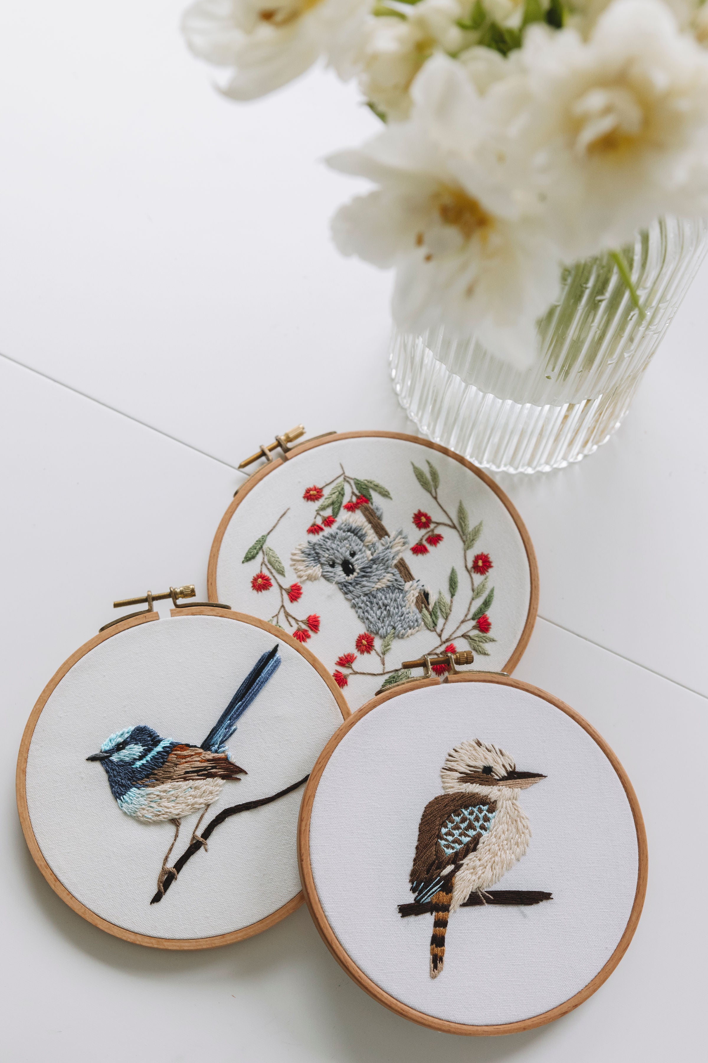 Three wooden embroidery hoops with detailed bird and koala designs arranged near a clear vase of white flowers on white surface