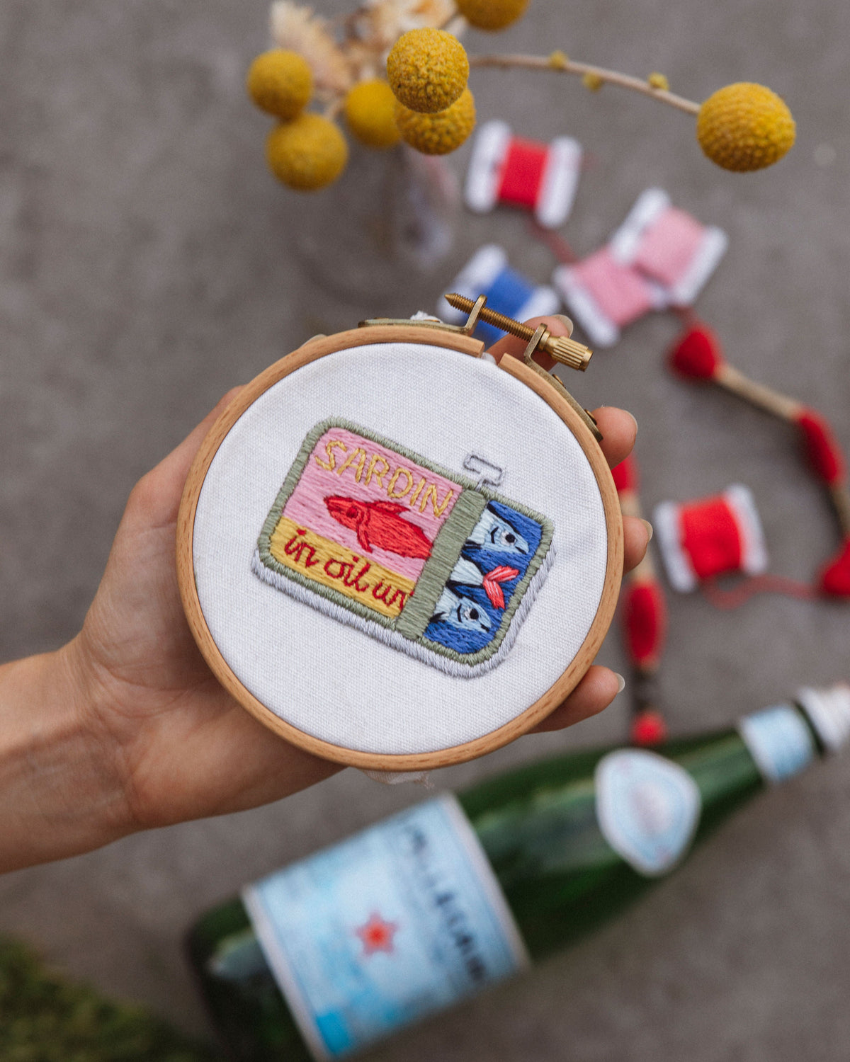 Embroidery hoop with colorful design held by a hand, with bottles and thread spools in the background.