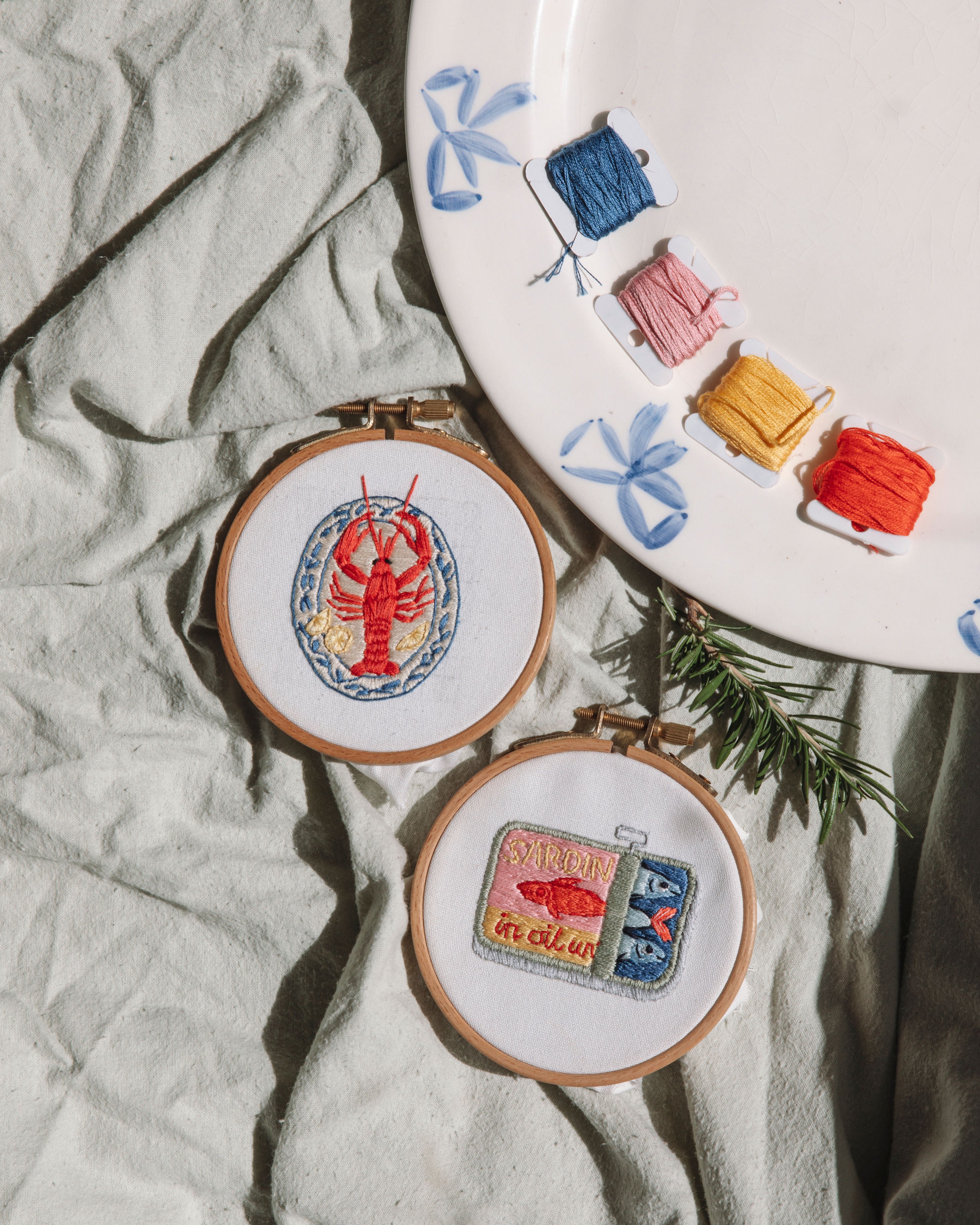 Mini Lobster embroidery hoop artwork with red crustacean design on white fabric, displayed with colorful thread spools and blue bows