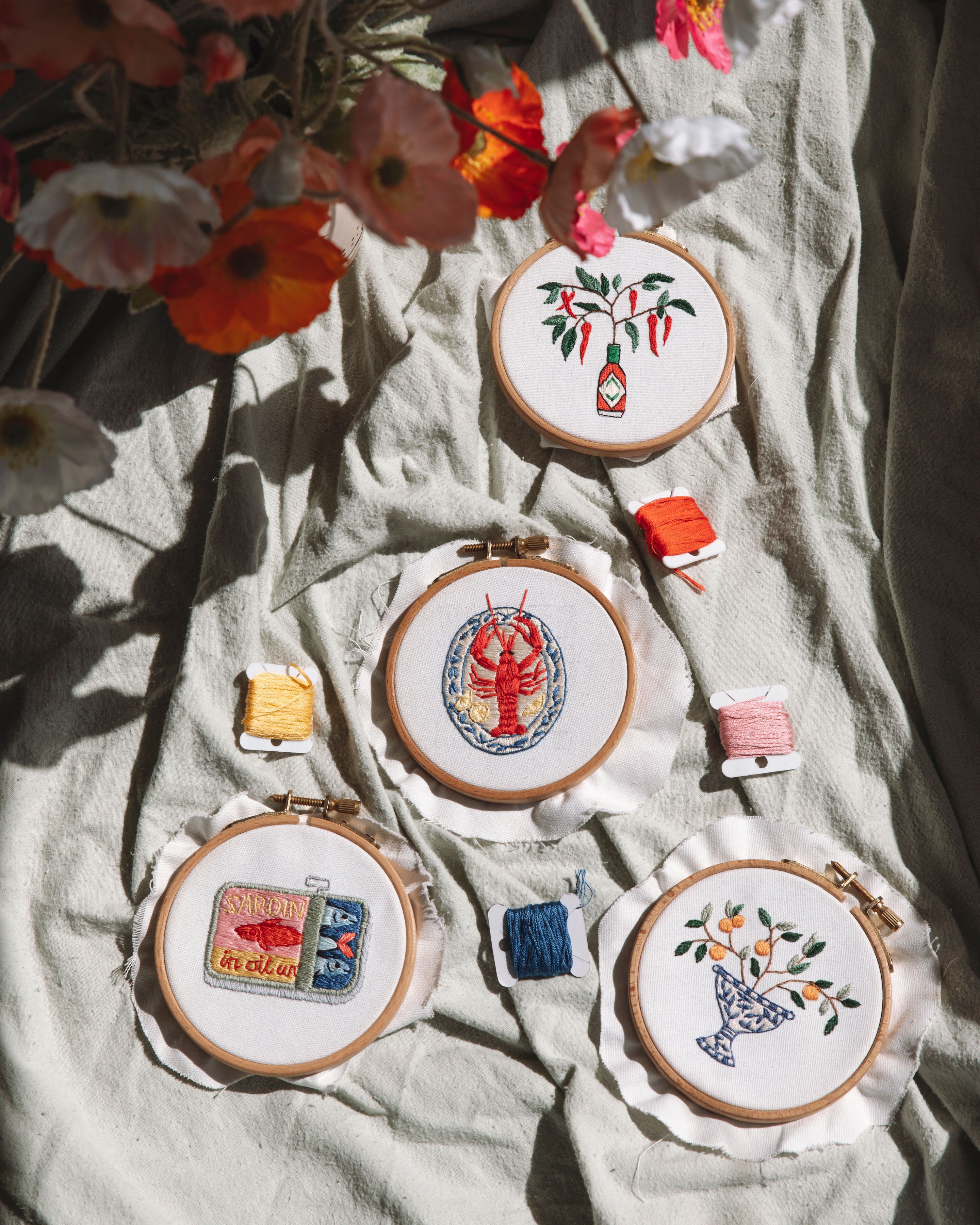 Four embroidery hoops with colorful stitched chili peppers, lobster, sardine tin, and fruit bowl designs on soft fabric with thread nearby
