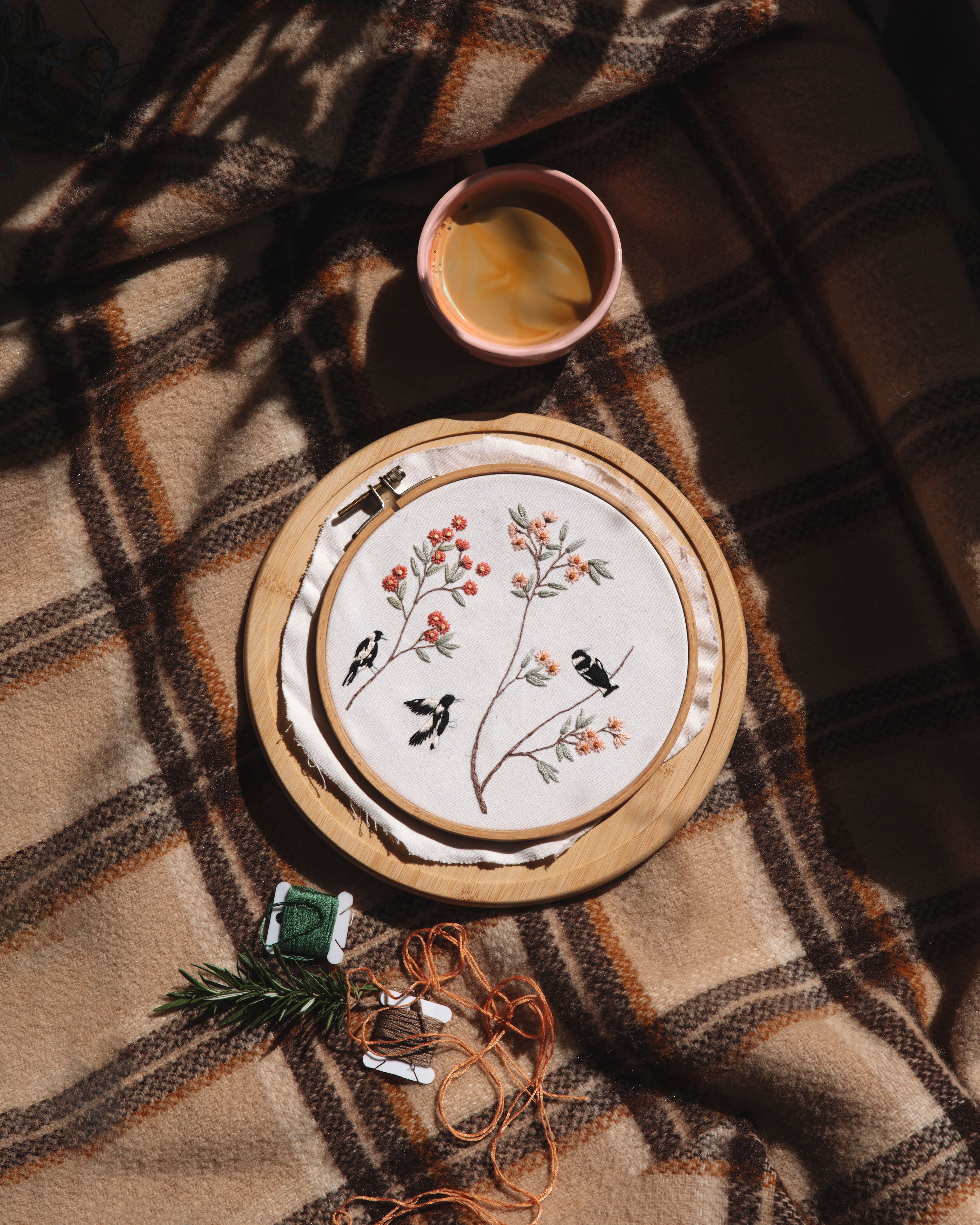 Spring Magpies embroidery hoop with floral and bird design displayed on a plaid blanket with thread and coffee cup.