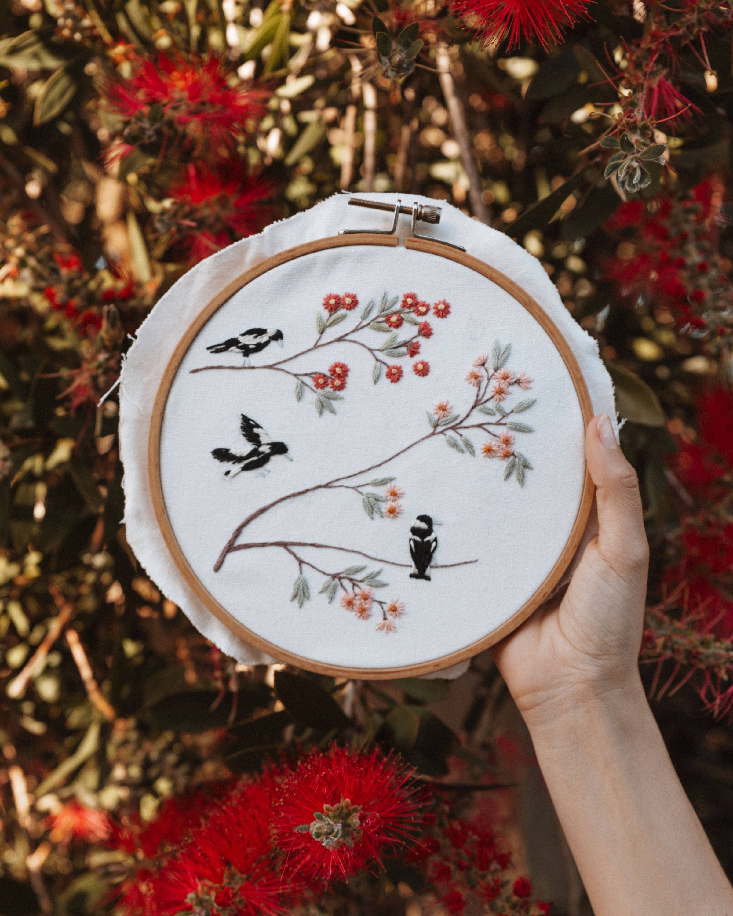 Hand holding embroidery hoop with Spring Magpies design featuring black birds and floral branches.