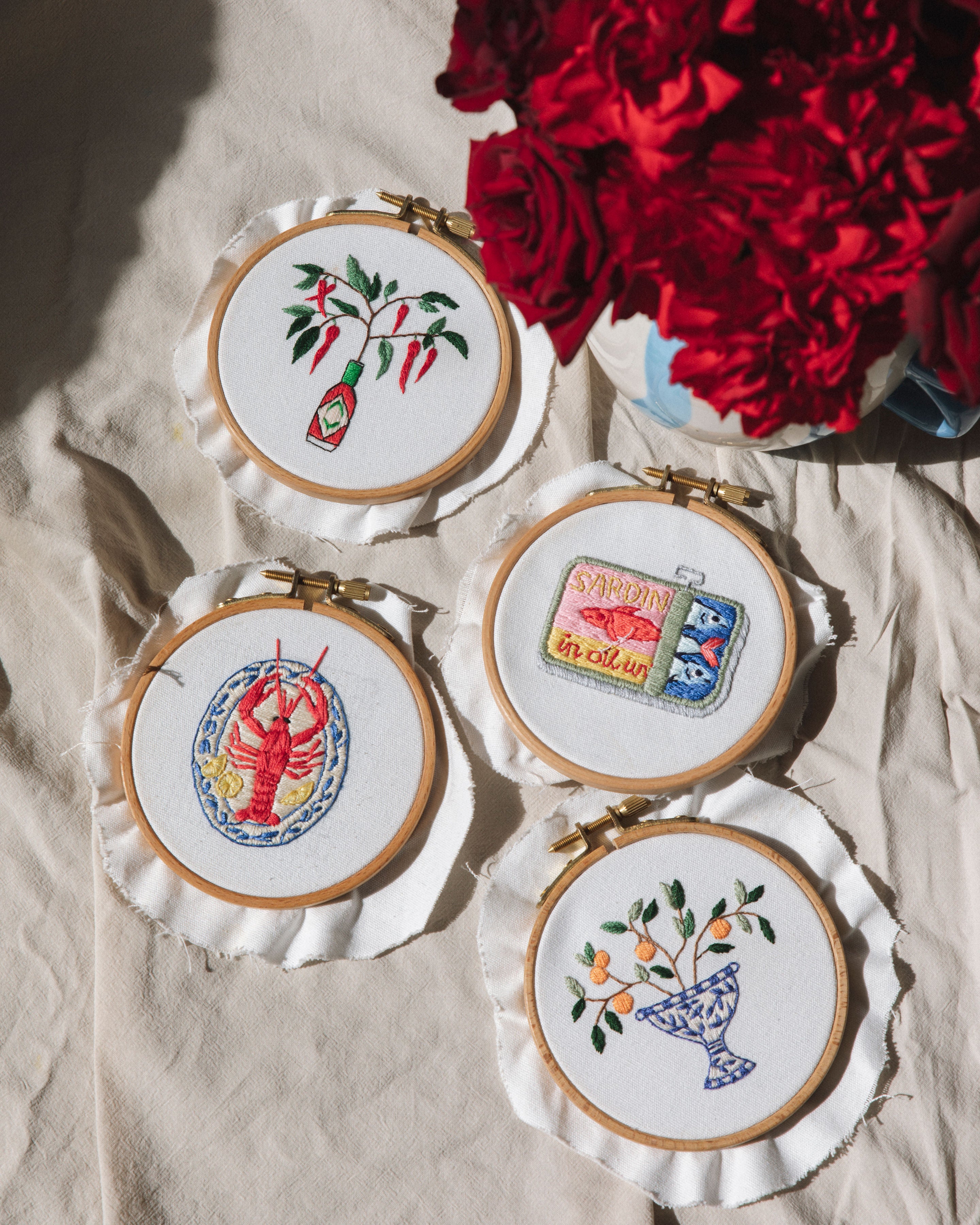 Mini Italian Vase embroidery hoops featuring whimsical designs of lobster, hot sauce bottle, sardine tin and floral arrangement on white fabric