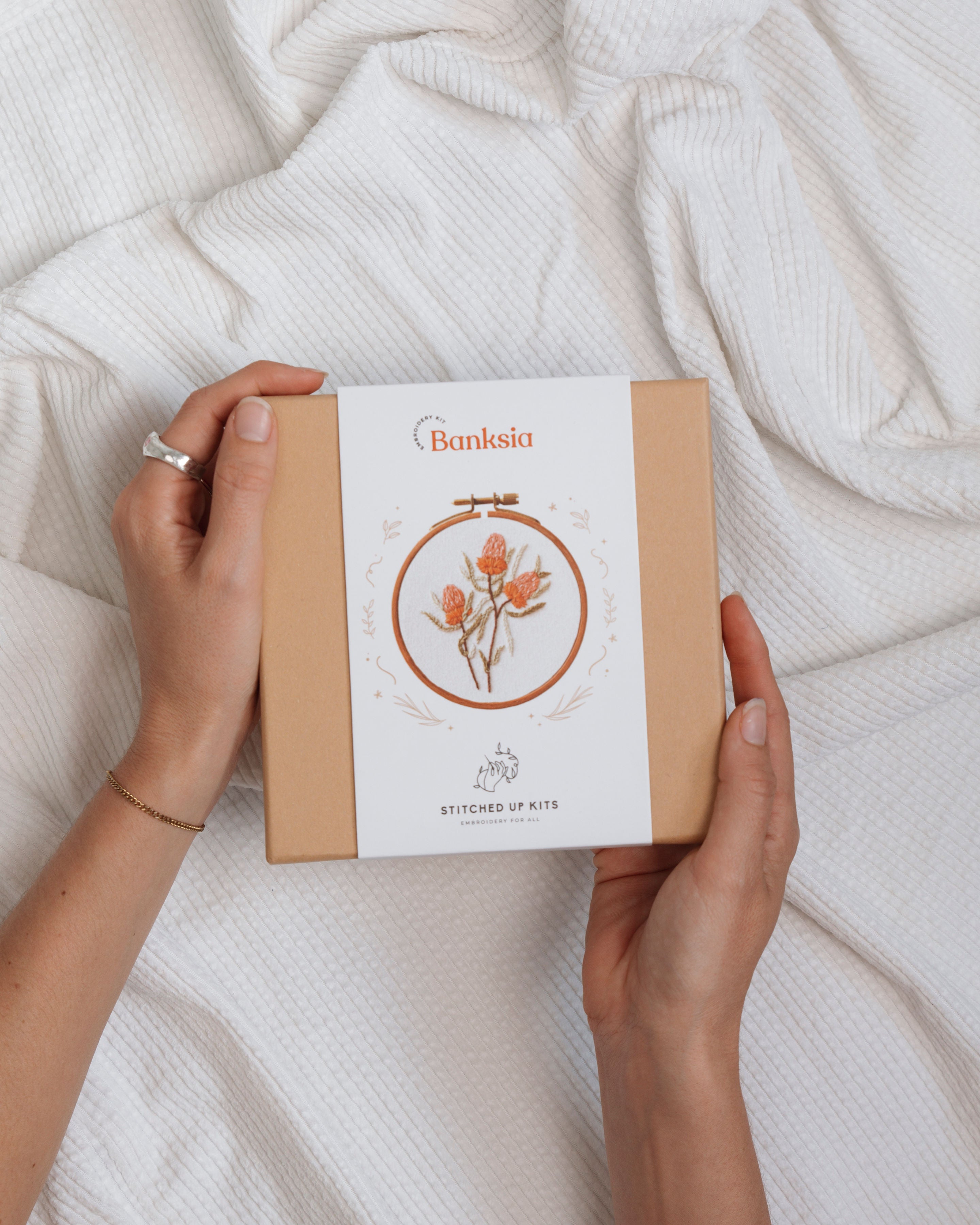All The Native Minis banksia embroidery kit box held over white textured fabric featuring orange flower design in circular hoop