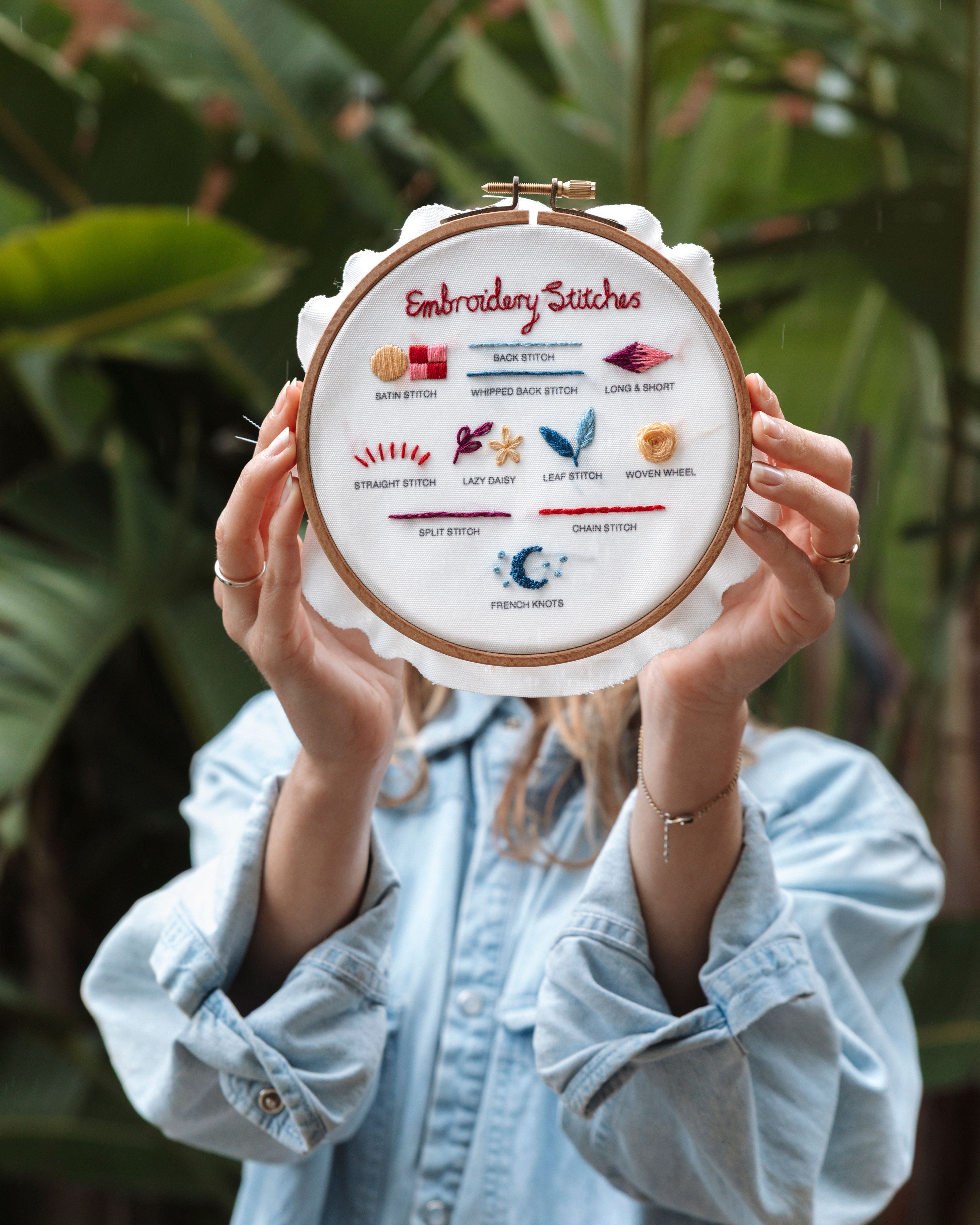 Person holding embroidery hoop showing colorful stitched embroidery stitch examples with labels on white fabric