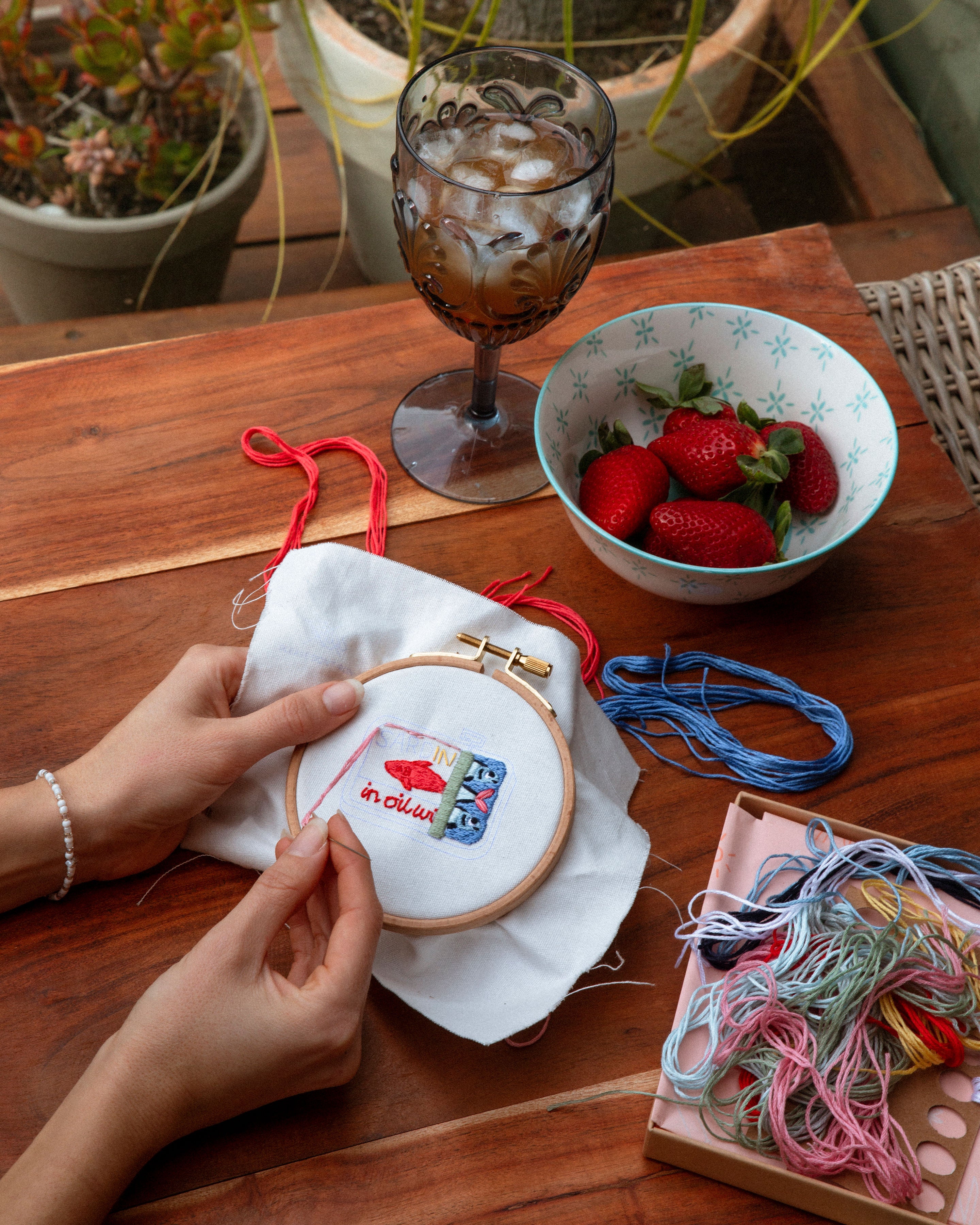 Hands working on a colorful embroidery hoop with threads, a bowl of strawberries, and a glass with ice on a wooden table