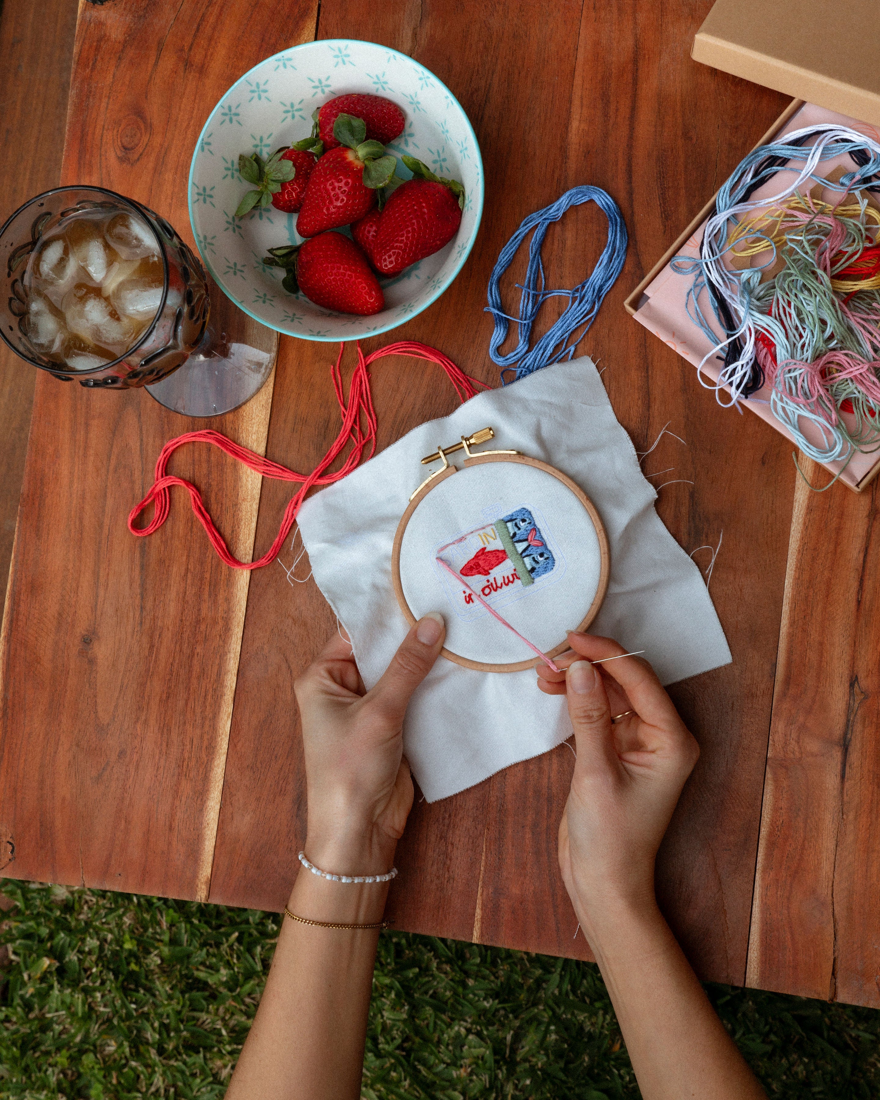 Person embroiderying on a wooden table with a bowl of strawberries and a drink.