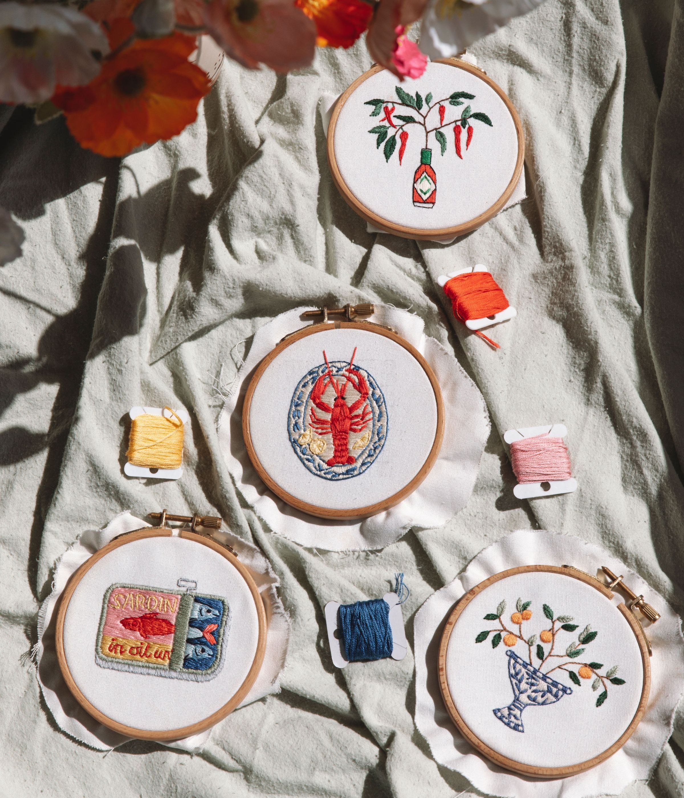Four wooden embroidery hoops showing a chili pepper bottle, lobster, sardine tins, and fruit in a blue vase with colorful thread nearby
