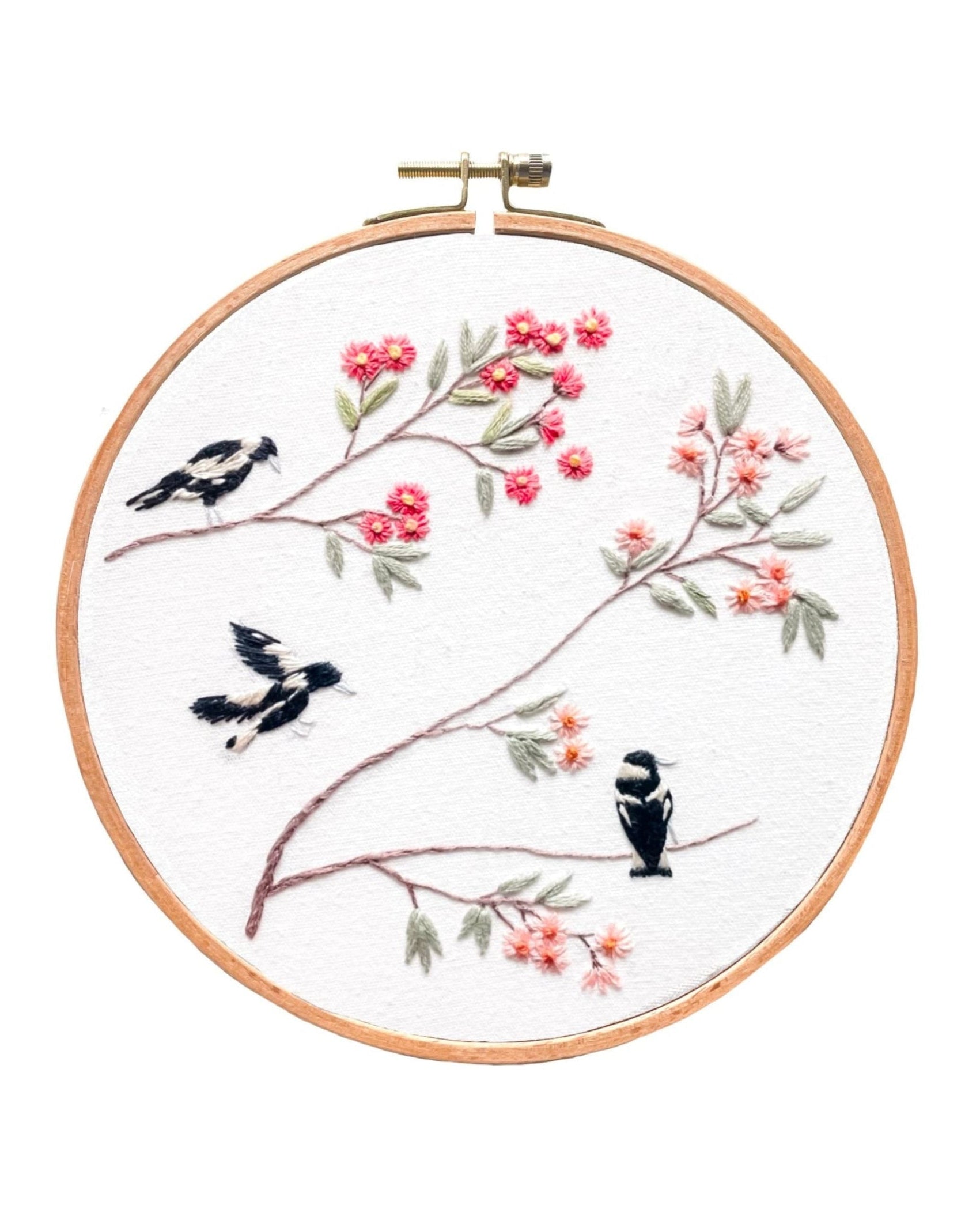 Spring Magpies embroidery hoop art with three magpies perched on branches with pink flowers on white fabric.