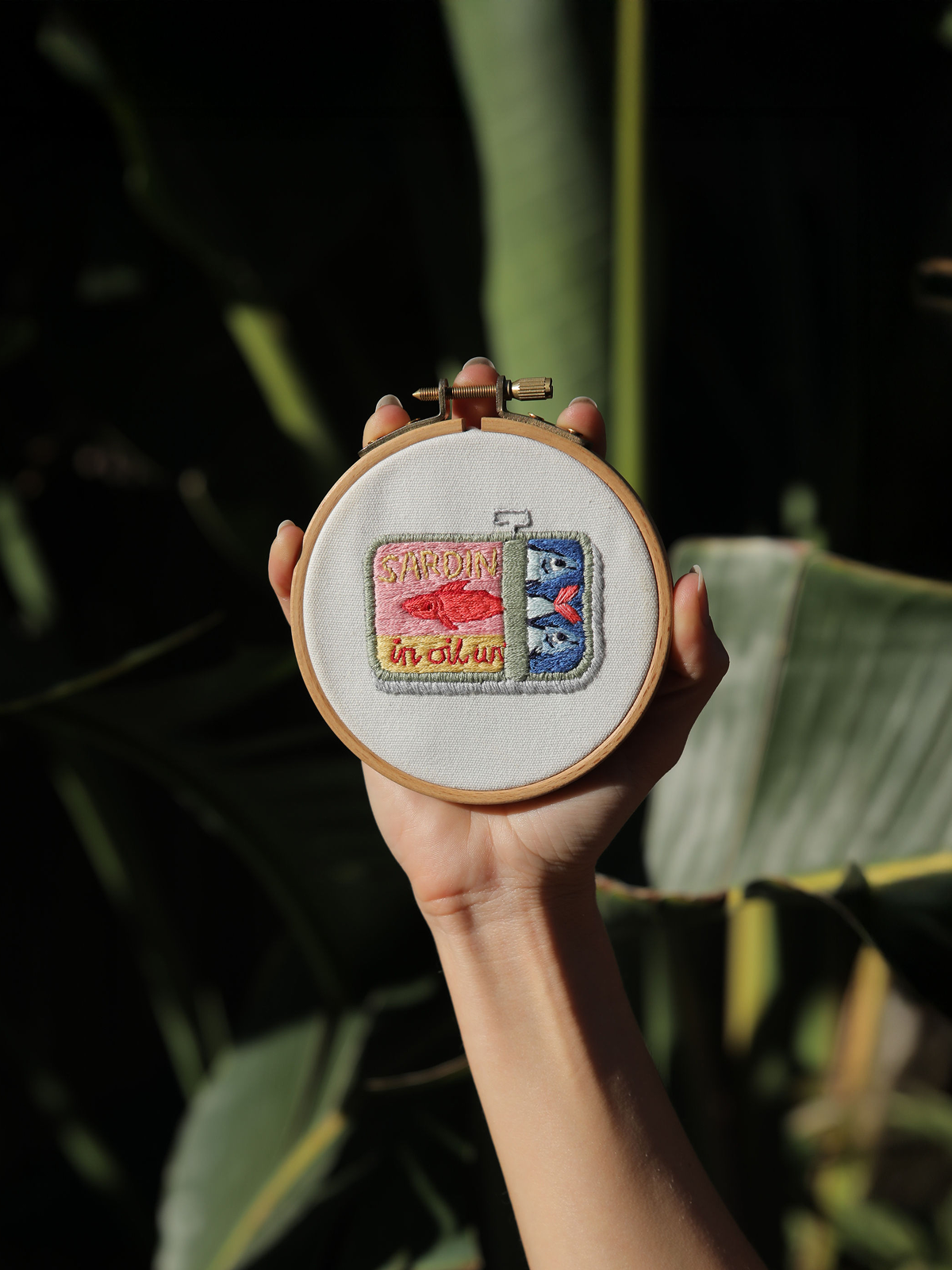 Hand holding embroidery hoop with a stitched design resembling a colorful opened sardine tin against a dark green leaf background