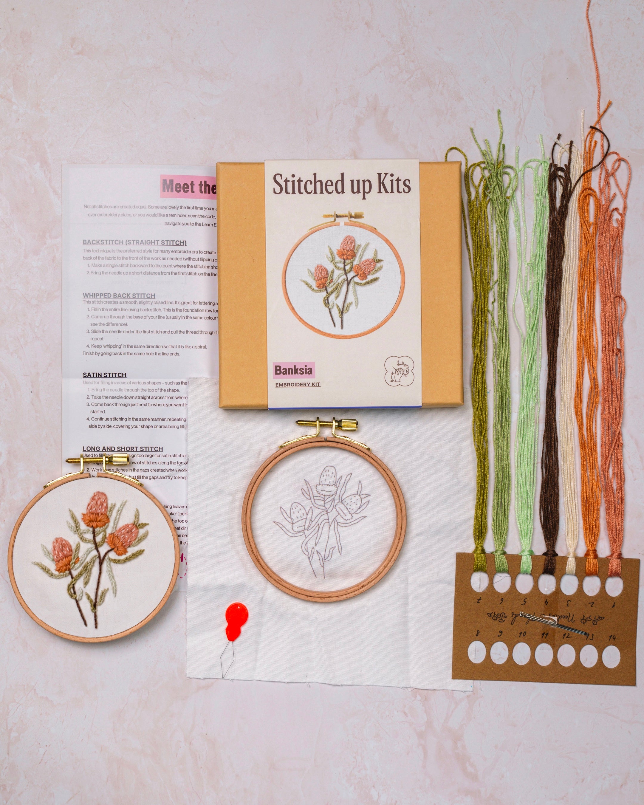 Embroidery kit with two completed hoop artworks, a box labeled 'Stitched up Kits', and colorful threads on a light background.