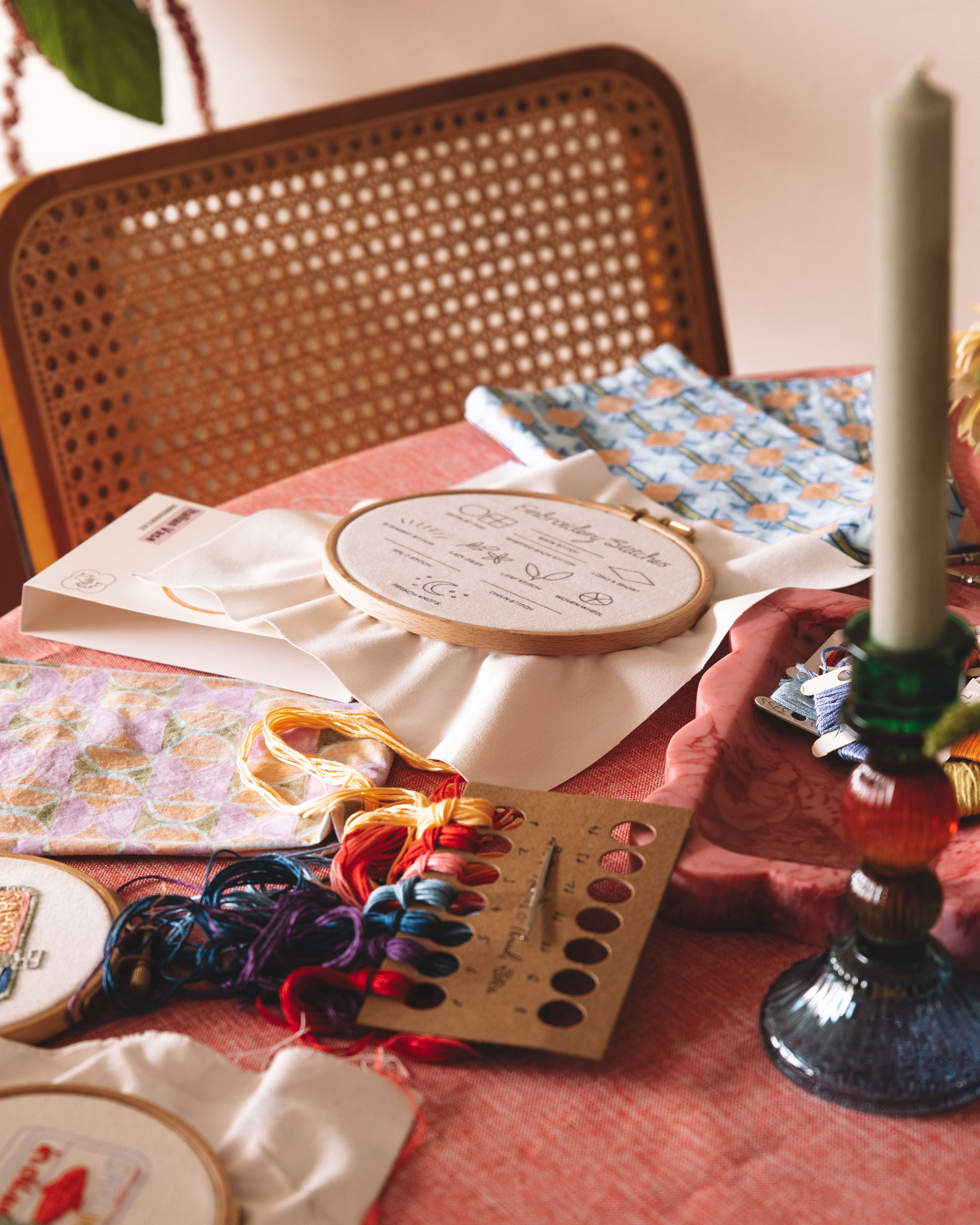 Embroidery hoop, scissors, and coloured threads on a table with a candle and chair in the background.