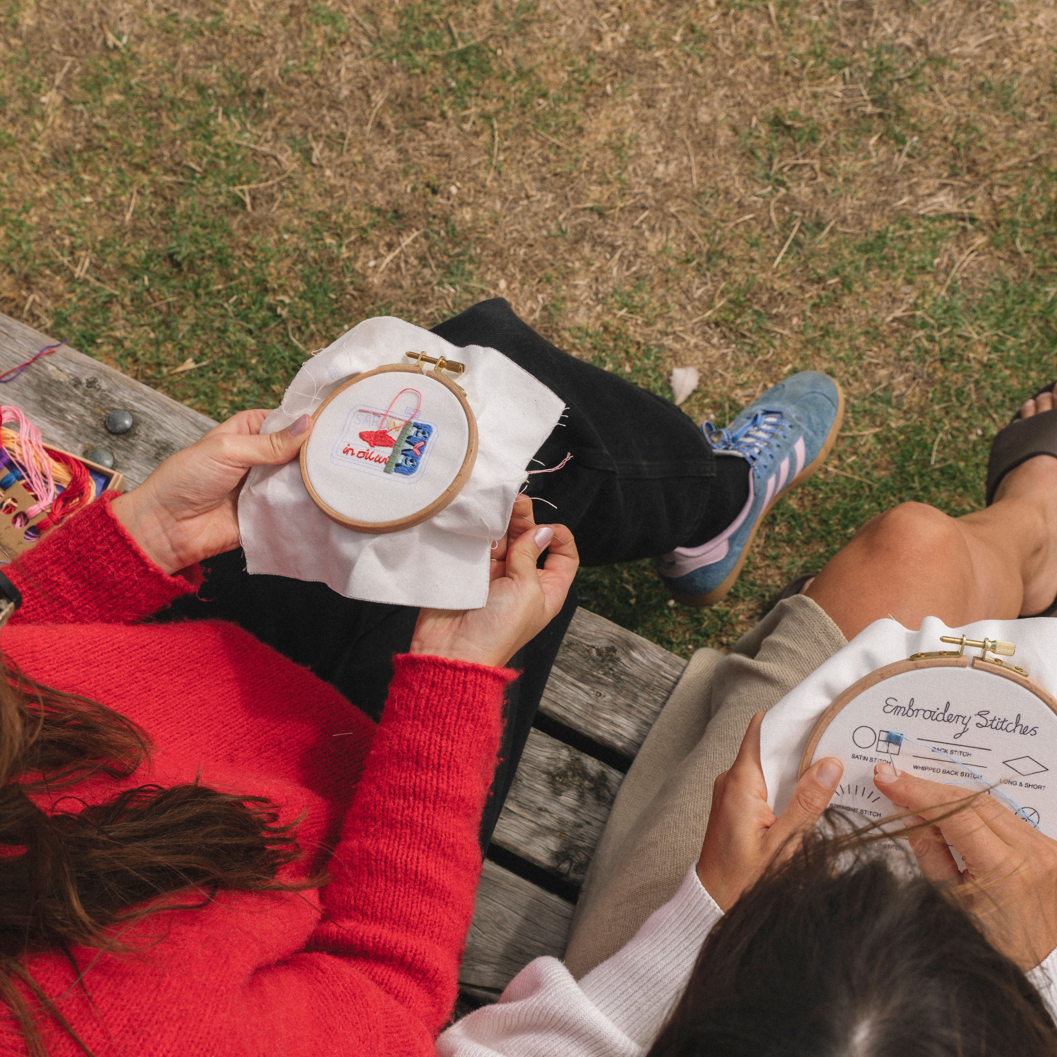 Two people sitting outdoors on a wooden bench, working on embroidery hoops with colorful thread details.
