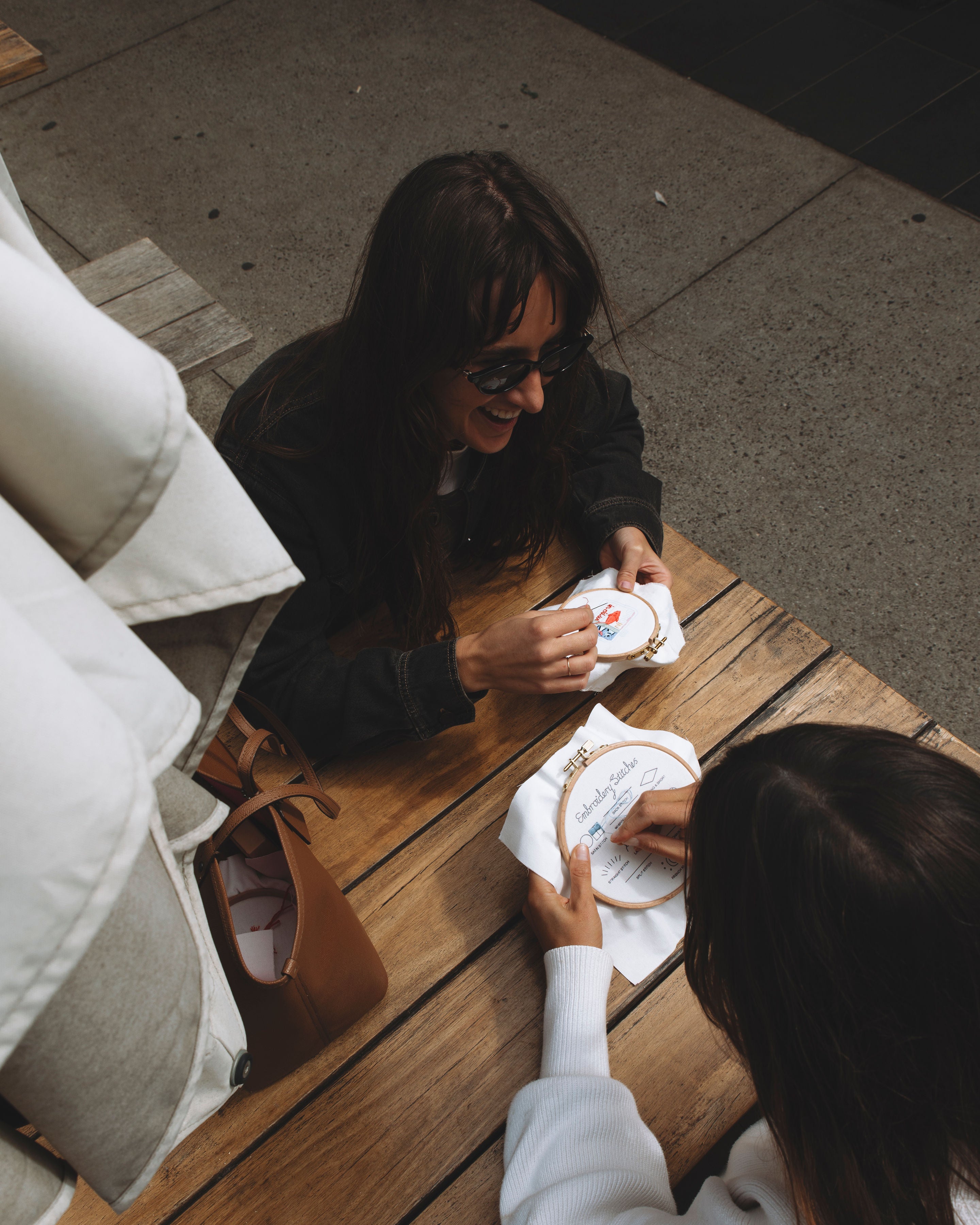 Two women sitting at a wooden table working on modern beginner embroidery kits with hoops and fabric outdoors