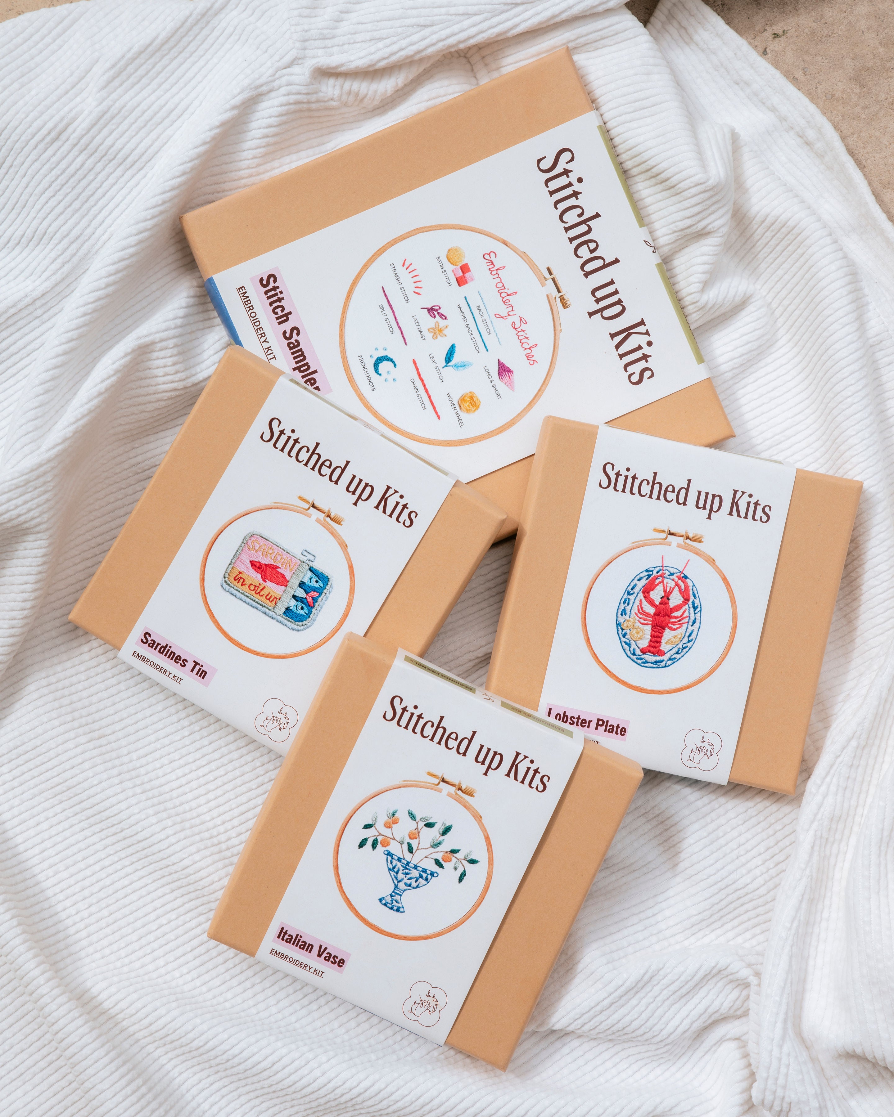 Four modern embroidery kits with stitched designs of a sardines tin, lobster, Italian vase, and stitch sampler on a white cloth