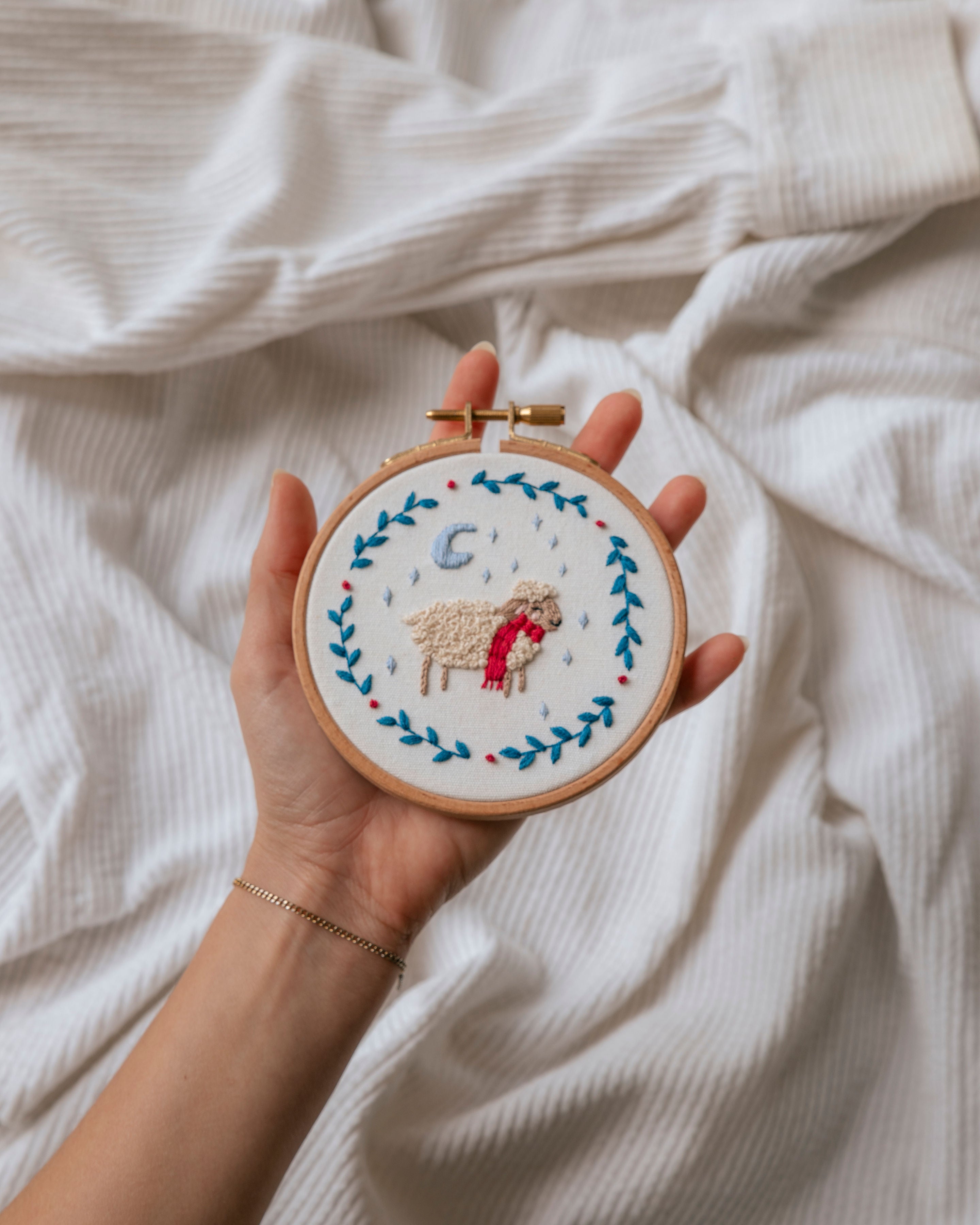 Hand holding an embroidery hoop with a stitched sheep wearing a red scarf surrounded by blue leaf vines on white fabric