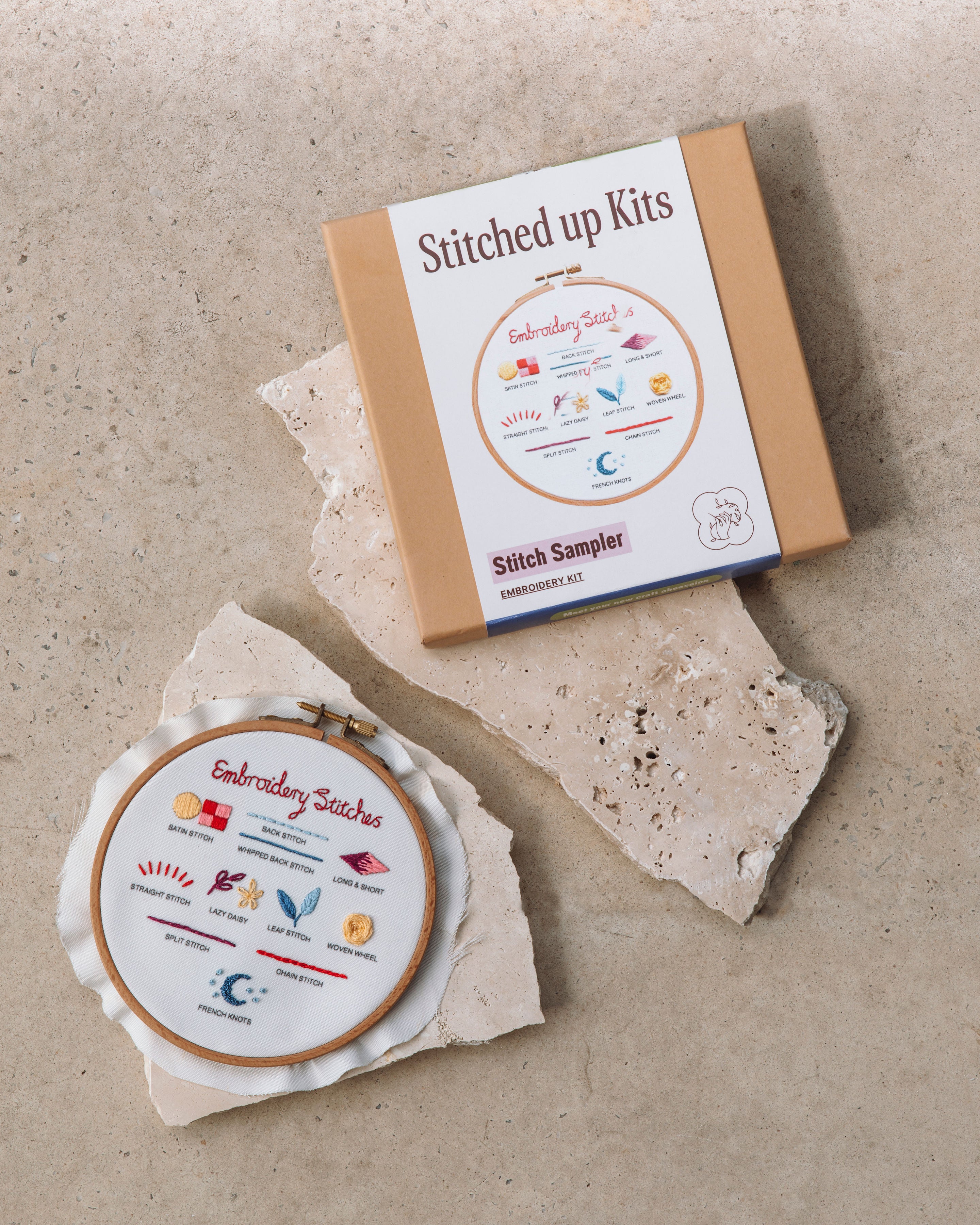 Embroidery hoop with colorful stitch sampler design and matching kit box on textured stone surface